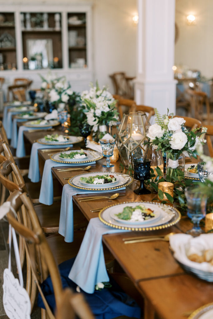 Salad plates are served on an elegant estate table with wooden farm tables.  Beautiful china, linens and glassware are color coordinated.  Floral arrangements with greenery and white flowers are set on the tables with candles.