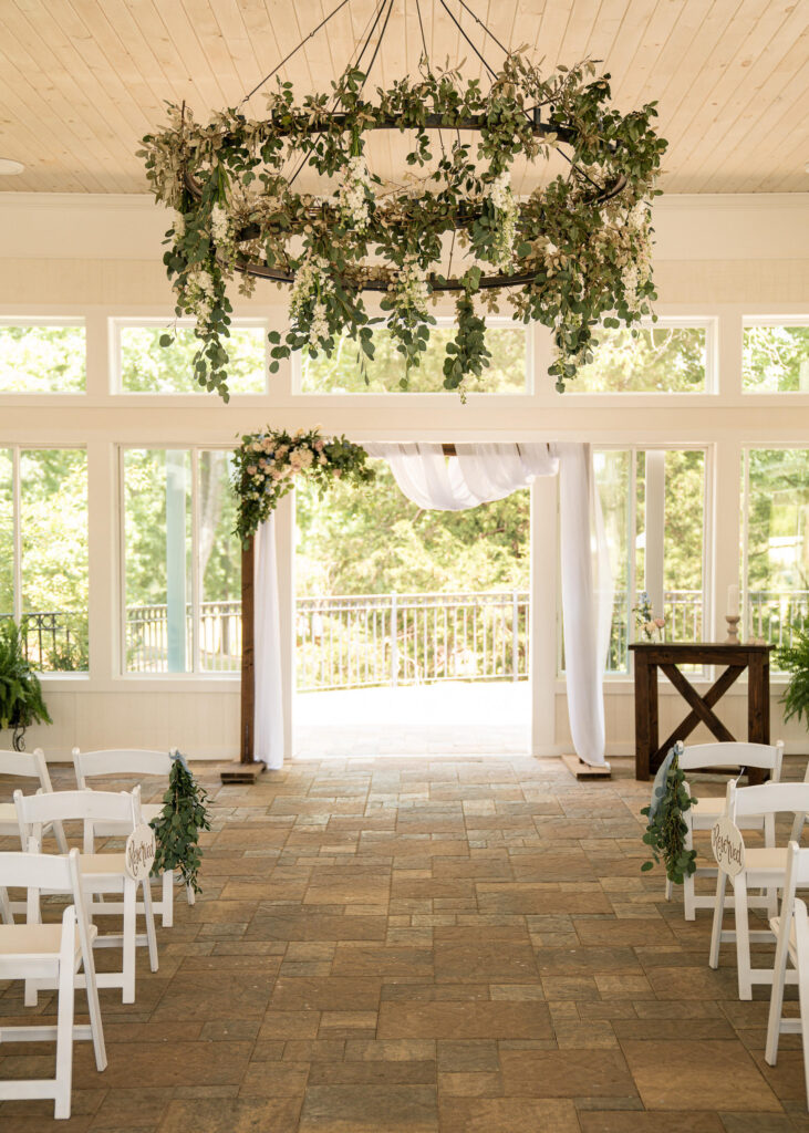 Greenery wrapped on double tiered wrought iron outdoor chandeliers and indoor ceremony site with wooden arbor adorned with draping and florals located on the covered and enclosed patio at a venue near Atlanta, GA 