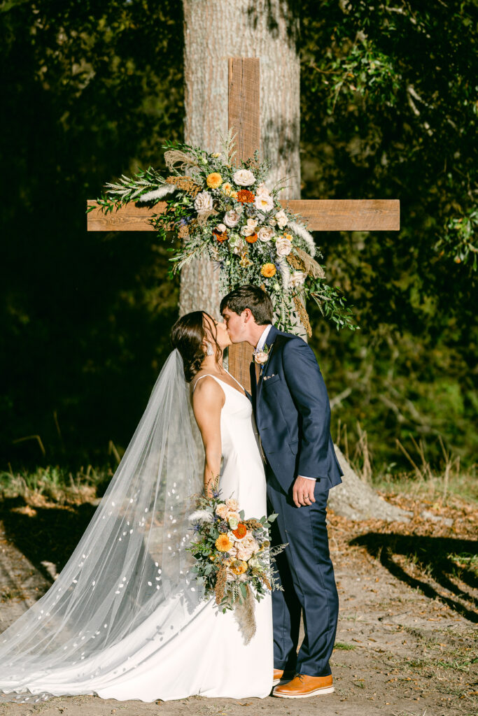 The beautiful bride holding her bridal bouquet and the handsome groom in his navy blue suit kiss at the outdoor ceremony site beneath the old wooden cross which holds a grand floral swag located at the Big Oak Tree ceremony at The Venue at Murphy Lane in Newnan, GA