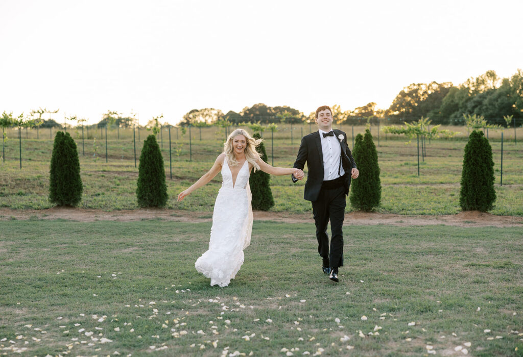 Bride in gorgeous wedding dress and groom in black tux running through the vineyard and outdoor ceremony site in a field at The Venue at Murphy Lane in Newnan, GA located one hour away from Atlanta, GA