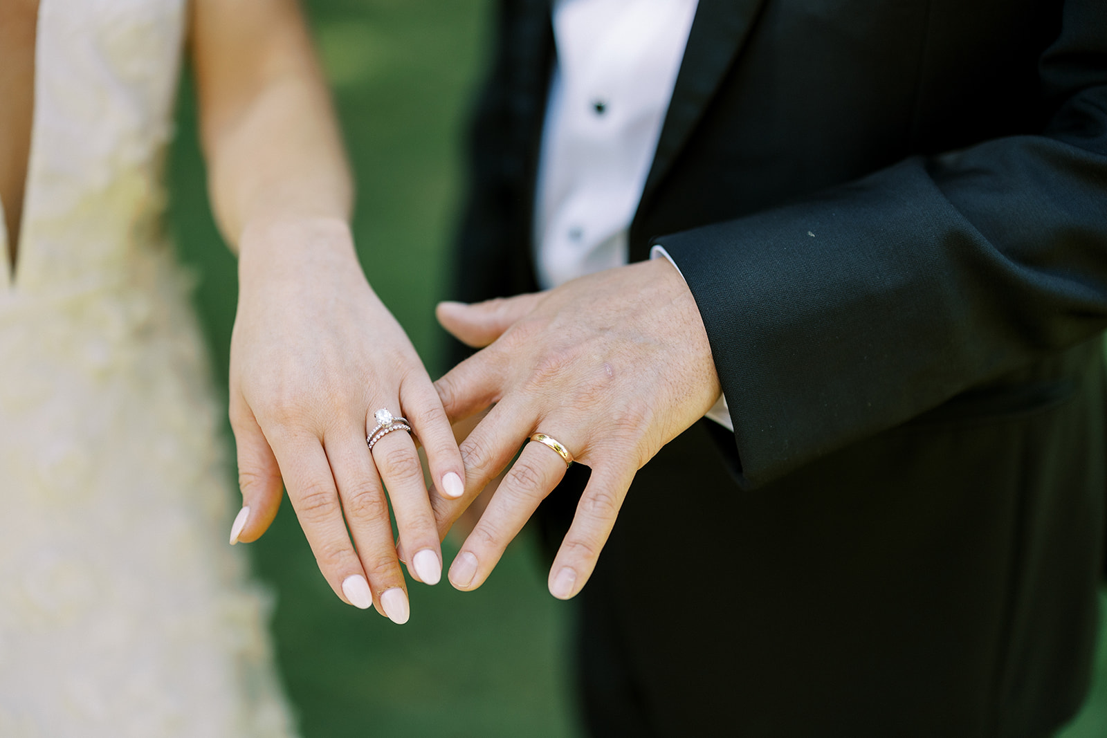 A bride and groom show their wedding rings on their hands which are touching after their wedding ceremony.  They are dressed for a wedding in a wedding gown and a black tuxedo.