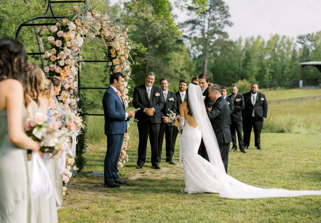 The bride and groom with the father of the bride and the officiant with the bridal party at the Big Oak Tree ceremony site with an oversized wrought iron garden style arbor adorned with spring florals at The Venue at Murphy Lane located in Newnan, GA