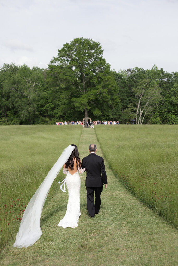 The father of the bride and the bride walk down the very long aisle through a hayfield on the farm which leads to the Big Oak Tree ceremony site located at a Newnan, GA wedding venue