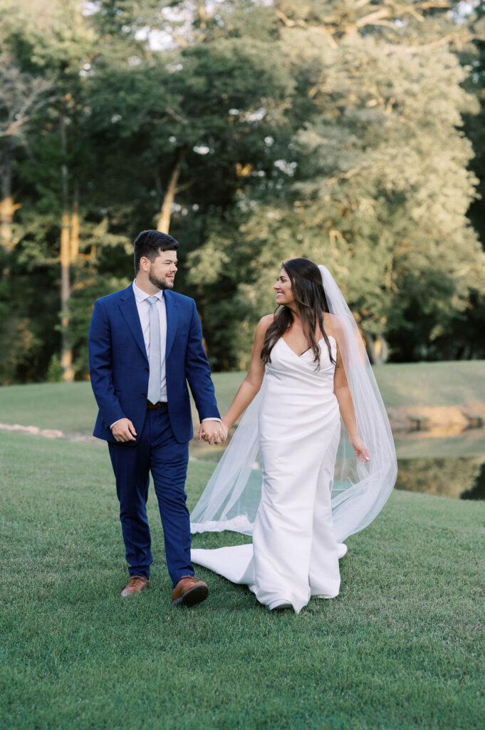 Bride in wedding dress and groom in navy suit walking along the pond on wedding day at a venue located in Newnan, GA