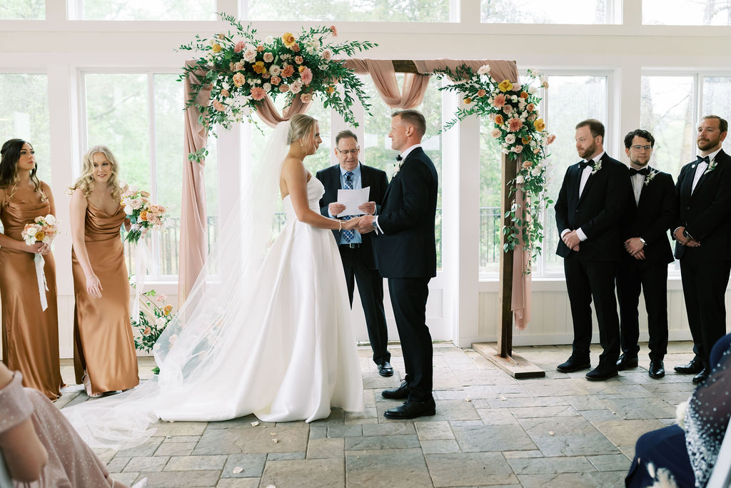 The bride and groom stand at a floral and draped arbor for an indoor wedding ceremony in a covered enclosed patio at a wedding venue located in Newnan, GA