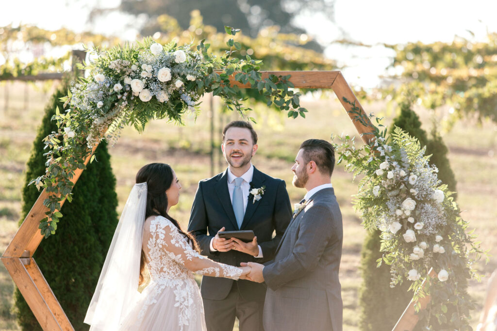 A wedding ceremony at an outdoor wedding venue in the midst of a vineyard and in front of a wooden arbor with ornate floral swags.  The officiant is speaking as the bride and groom hold hands and face each other.