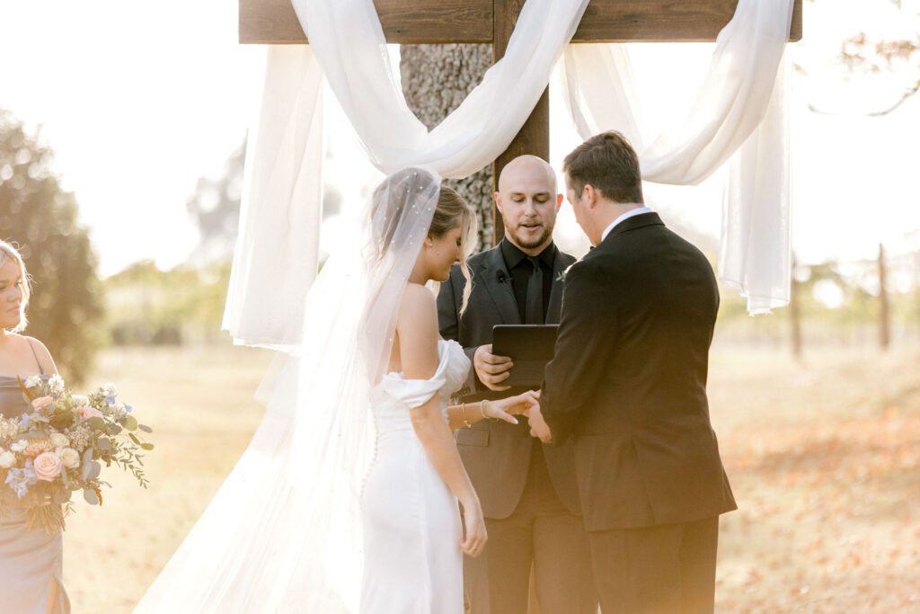 A groom in a black suit is placing a wedding ring on the finger of a bride who is wearing a white wedding gown during an outdoor wedding ceremony.  An officiant is speaking to the couple as they all stand in front of a large wooden cross which has white draping.  
The maid of honor holds the brides floral bouquet.