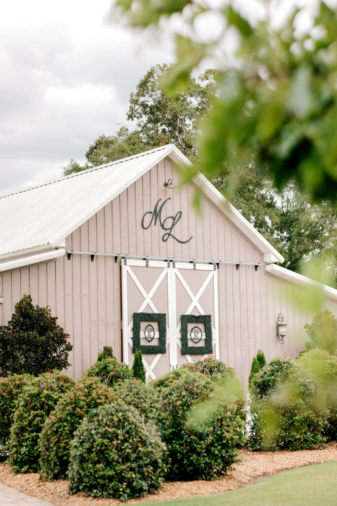 A wedding venue barn in the Atlanta area with the large barn doors closed.  Shrubs and trees are part of the landscaping in front of the building.  There are large trees behind the barn.