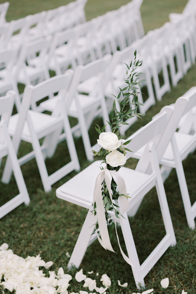 White padded resin chairs with aisle markers created with  white roses, greenery and white ribbons are all set up for a wedding ceremony in the valley at The Venue at Murphy Lane located in Newnan, GA 