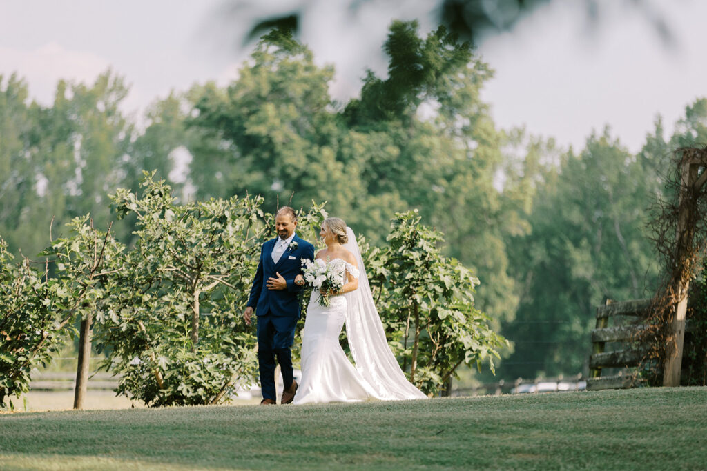 Beautiful bride in her wedding dress with long veil is escorted down the aisle to the valley wedding site by her stepfather passing by the fig trees and beautifully manicured green grass in Newnan, GA at The Venue at Murphy Lane