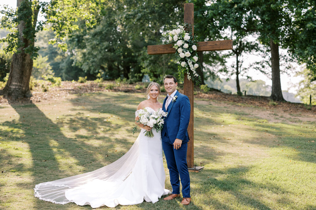 Bride in her long white wedding and gorgeous floral bouquet and groom in his navy blue suit stand beneath the the 9 1/2 ft wooden cross adorned with a large asymmetrical floral swag with white flowers and greenery which stands at the altar at the ceremony site in the valley at The Venue at Murphy Lane located in Newnan, GA