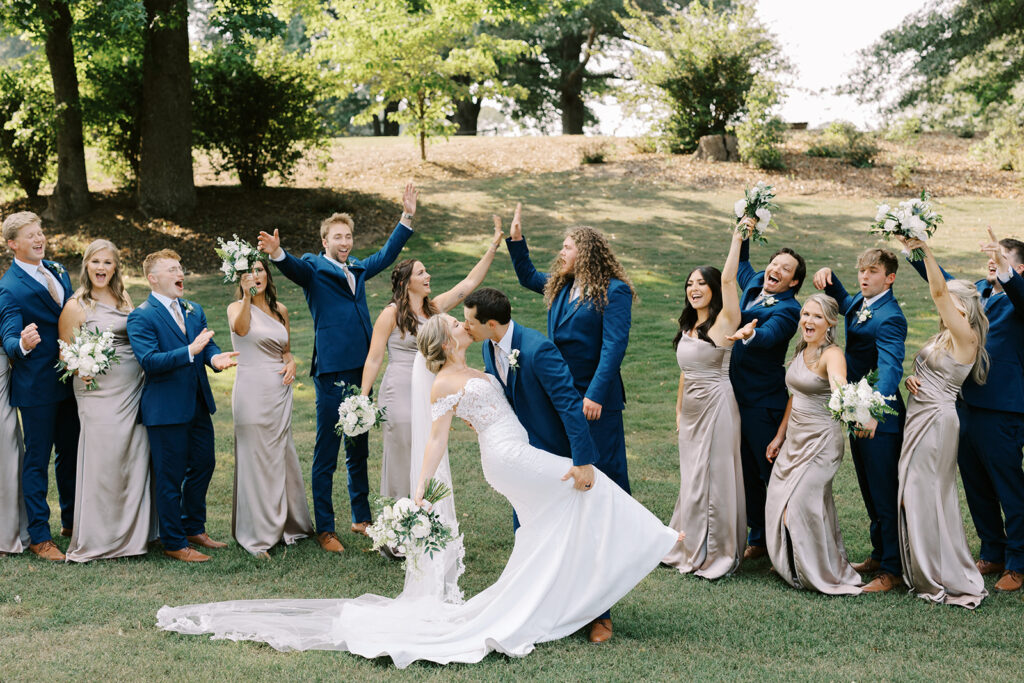 The bride and groom kiss after the wedding ceremony in the valley surrounded by their bridal party of groomsmen in navy blue suits and bridesmaids in champagne bridesmaid dresses and lovely floral bouquets at The Venue at Murphy Lane located in Newnan, GA just on hour away from Atlanta, GA